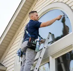 Professional window cleaner on a ladder using a squeegee to clean an exterior arched window, Boulder, CO.